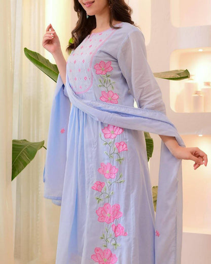 Woman wearing a light blue traditional outfit with pink floral embroidery in a bright setting.