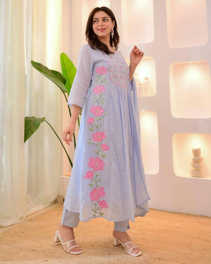 Woman wearing a light blue dress with pink floral embroidery in a bright room.